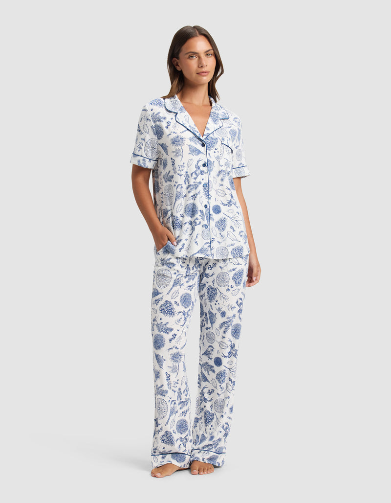 A woman stands barefoot in the Cozy Earth Women's Bamboo Stretch-Knit Short Sleeve Pajama Set, featuring a blue and white botanical print with a collared button-up top and loose pants. She has one hand in her pocket and looks relaxed.