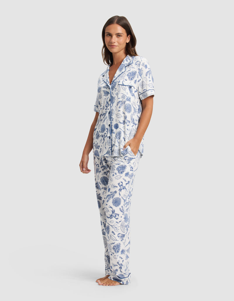 A woman stands barefoot against a light background, wearing Cozy Earth's Women's Bamboo Stretch-Knit Short Sleeve Pajama Set in a blue and white floral pattern with a button-up top and long pants.