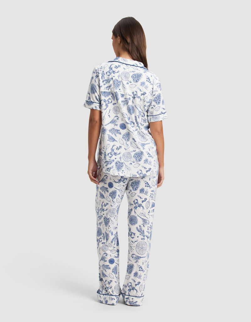 A woman with long brown hair faces away, wearing the Cozy Earth Women's Bamboo Stretch-Knit Short Sleeve Pajama Set in a blue and white botanical pattern with navy trim, set against a plain light gray background.