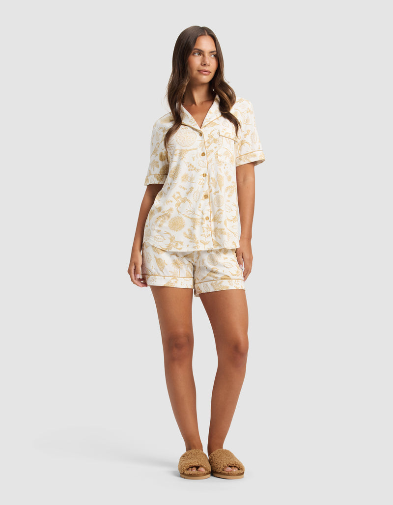 A woman with long brown hair wears the Cozy Earth Women's Bamboo Stretch-Knit Short Sleeve Pajama Set featuring a light floral pattern, paired with brown slippers, and stands against a plain background.