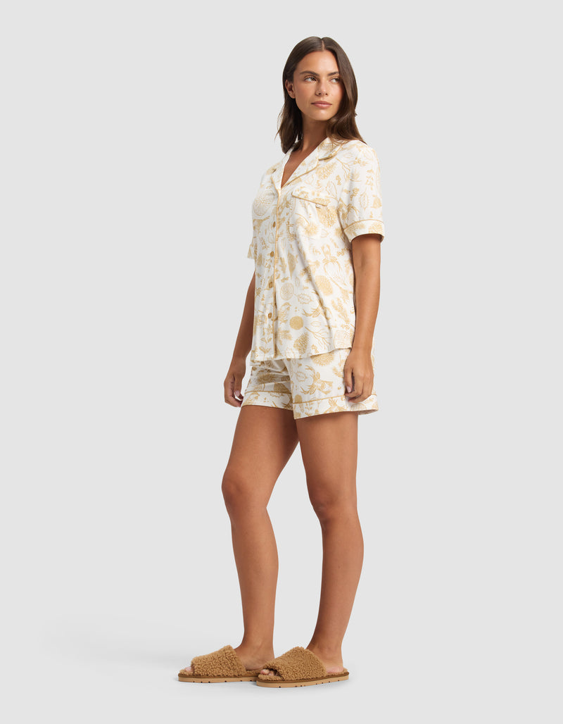 A woman stands looking to the side, wearing Cozy Earth’s Women's Bamboo Stretch-Knit Short Sleeve Pajama Set in a beige botanical print, paired with brown textured slippers. The background is plain light gray.