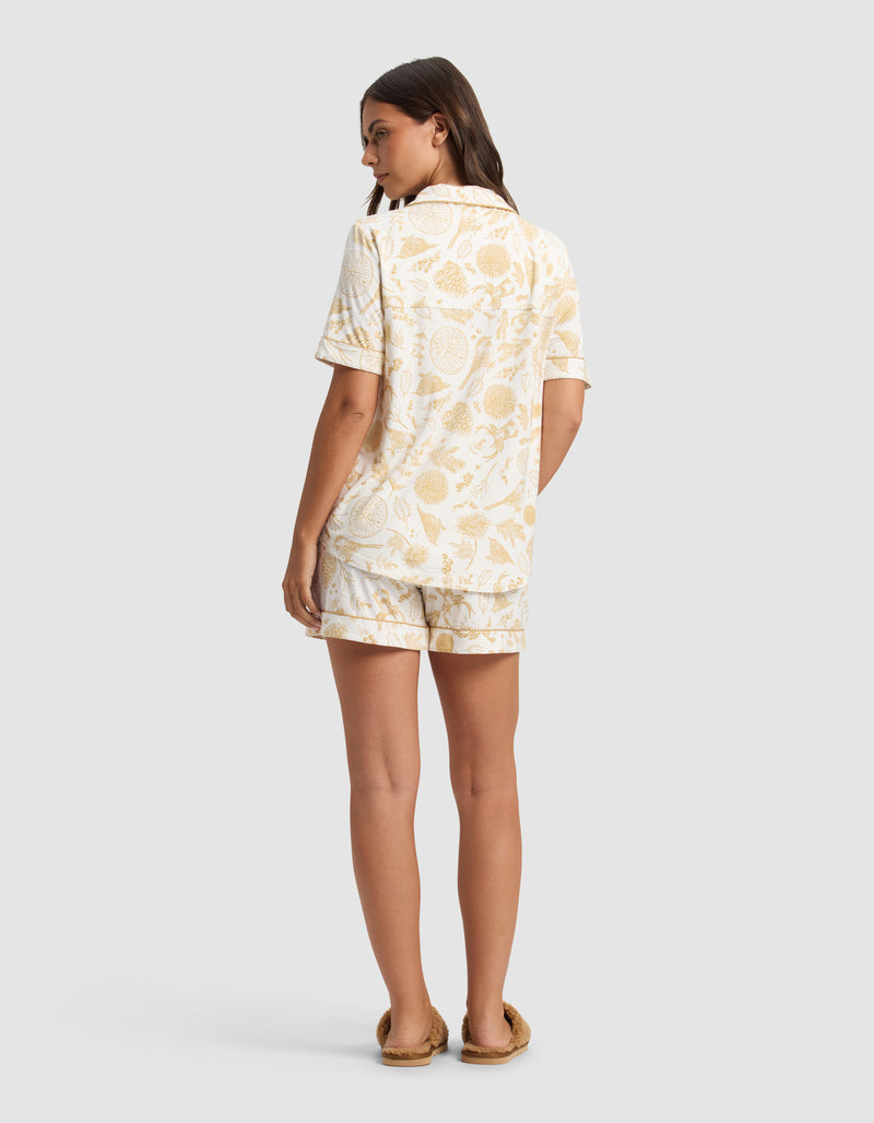 A woman faces away, wearing the Cozy Earth Women's Bamboo Stretch-Knit Short Sleeve Pajama Set in a light yellow botanical print and slippers, set against a plain light gray background.