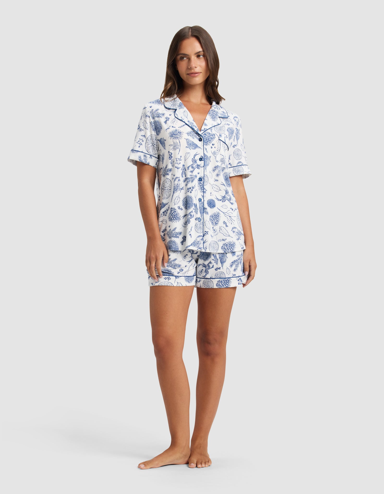 A woman poses barefoot against a light gray backdrop, wearing the Cozy Earth Women's Bamboo Stretch-Knit Short Sleeve Pajama Set—a matching button-up top with a collar and shorts, featuring a blue botanical print on white.