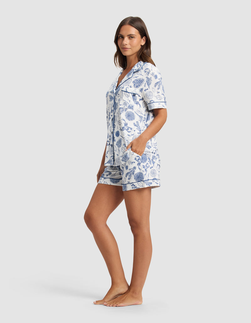 A woman stands barefoot, looking relaxed against a plain light background, wearing the Cozy Earth Women's Bamboo Stretch-Knit Short Sleeve Pajama Set with blue and white floral patterns and her hands in the shorts' pockets.
