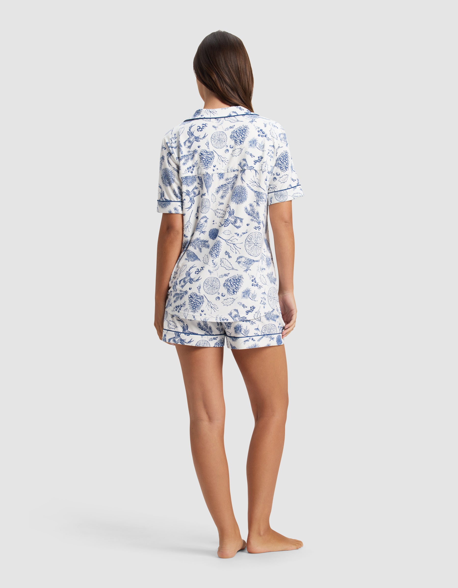 A woman stands barefoot, facing away, wearing the Cozy Earth Women's Bamboo Stretch-Knit Short Sleeve Pajama Set with a blue and white botanical print. The background is plain light gray.|Color:Winter Toile Pacific
