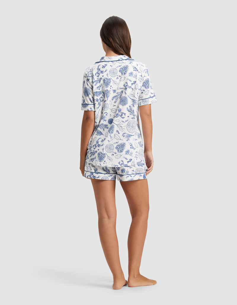 A woman stands barefoot, facing away, wearing the Cozy Earth Women's Bamboo Stretch-Knit Short Sleeve Pajama Set with a blue and white botanical print. The background is plain light gray.