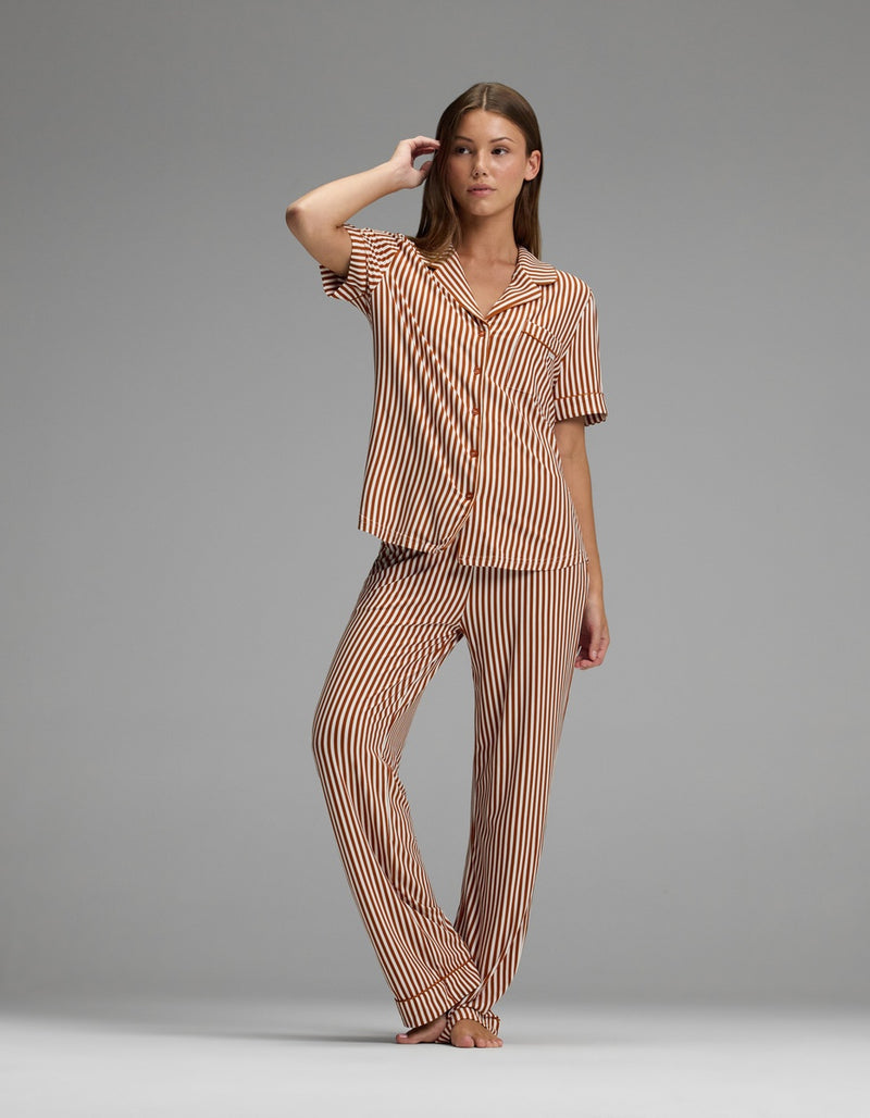 A woman stands barefoot against a plain gray background, wearing the Cozy Earth Women’s Bamboo Stretch-Knit Short Sleeve & Pant Pajama Set in matching brown and white vertical stripes.