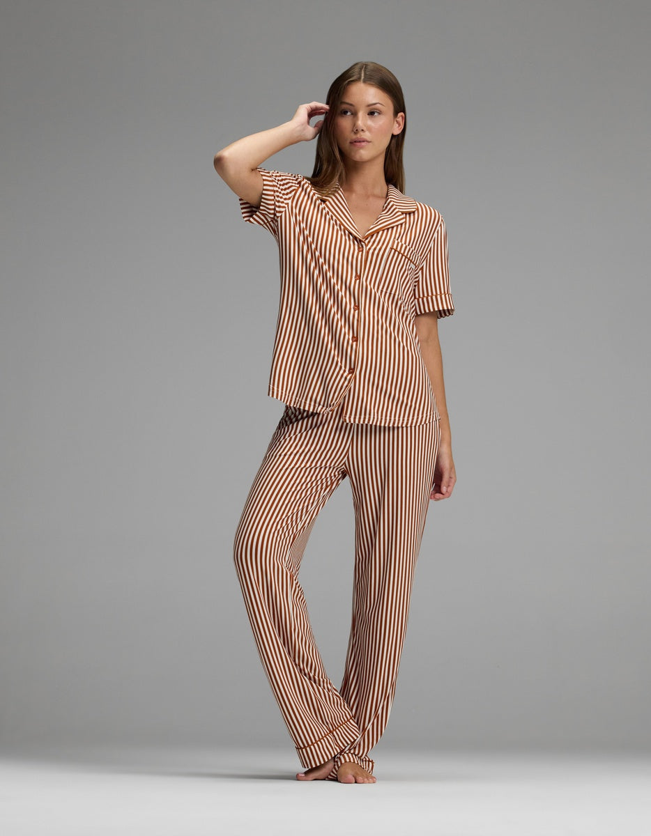 A woman stands barefoot against a plain gray background, wearing the Cozy Earth Women’s Bamboo Stretch-Knit Short Sleeve & Pant Pajama Set in matching brown and white vertical stripes.|Color:Market Stripe in Cinnamon
