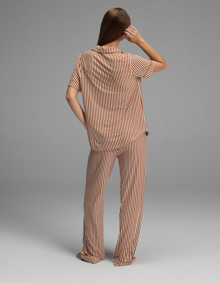 A person stands facing away, wearing the Cozy Earth Women’s Bamboo Stretch-Knit Short Sleeve & Pant Pajama Set in a loose fit with tan and white vertical stripes. They have long, straight brown hair and hold one arm bent at the elbow, touching the side of their head. The background is plain gray.