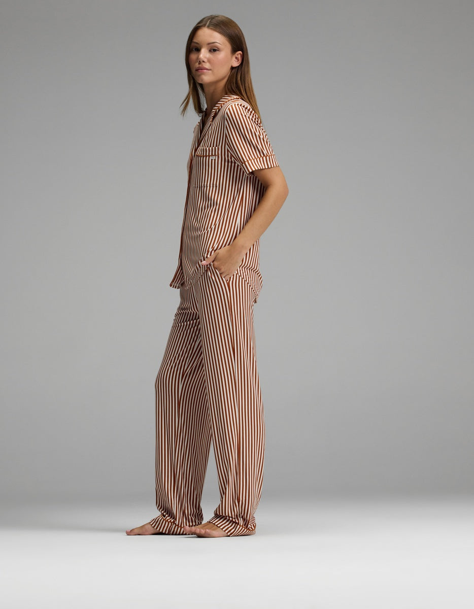 A person stands barefoot, facing sideways with hands in pockets, wearing Cozy Earth’s Women’s Bamboo Stretch-Knit Short Sleeve & Pant Pajama Set in a striped pattern against a plain gray background.