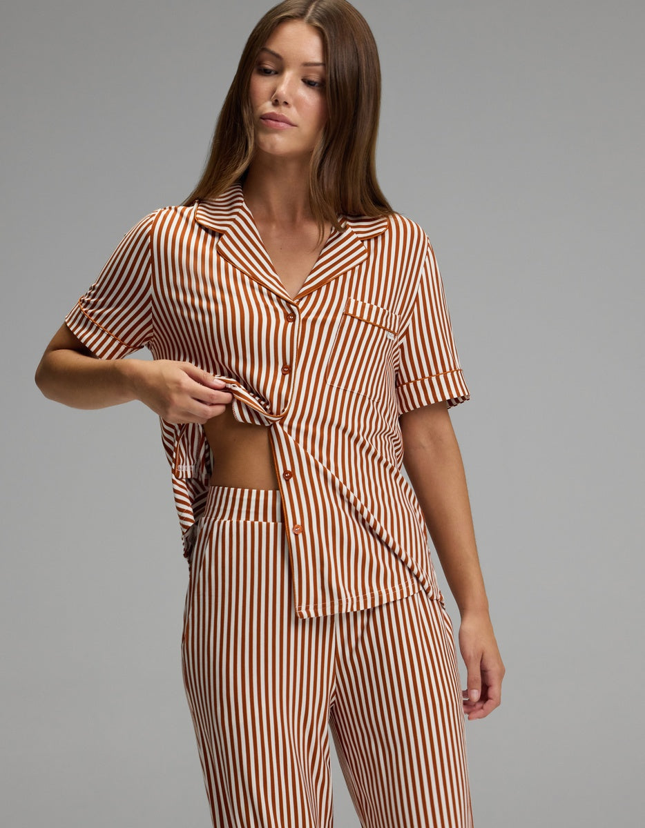 A woman with long brown hair stands against a plain grey background, lifting part of her shirt to reveal her side while wearing the Cozy Earth Women’s Bamboo Stretch-Knit Short Sleeve & Pant Pajama Set in a brown and white vertical stripe pattern.