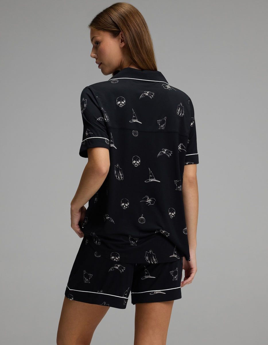 A person with light brown hair wears the Cozy Earth Women’s Bamboo Stretch-Knit Short Sleeve Pajama Set in black, featuring white piping and small skull, bat, cat, and moon designs, while standing against a plain gray background.