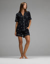 A woman stands barefoot against a plain gray background, wearing the Cozy Earth Women’s Bamboo Stretch-Knit Short Sleeve Pajama Set in black with white piping and small white designs, featuring a short-sleeve button-up top and matching shorts.|Color:Halloween Print
