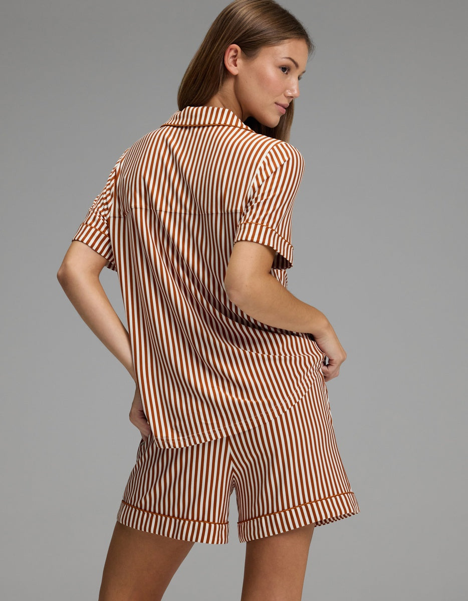A woman stands sideways, looking over her shoulder, wearing the Cozy Earth Women’s Bamboo Stretch-Knit Short Sleeve Pajama Set in brown and beige stripes against a plain gray background.