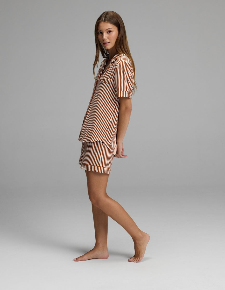 A woman stands barefoot on a gray background, wearing the Cozy Earth Women’s Bamboo Stretch-Knit Short Sleeve Pajama Set in a brown-and-white striped pattern with shorts. She is turned slightly to the side and looking toward the camera.