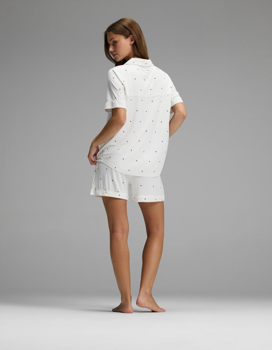 A woman stands barefoot facing away, wearing the Cozy Earth Women’s Bamboo Stretch-Knit Short Sleeve Pajama Set—a loose white shirt and matching shorts with a subtle dot pattern—against a plain gray background.
