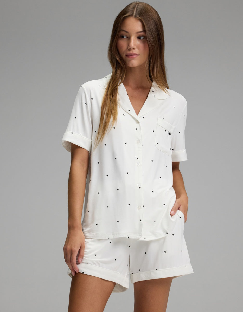 A woman stands wearing the Cozy Earth Women’s Bamboo Stretch-Knit Short Sleeve Pajama Set, a matching white ensemble with short sleeves and shorts adorned with small black polka dots. She has long straight hair and is looking to the side, with one hand in her pocket.