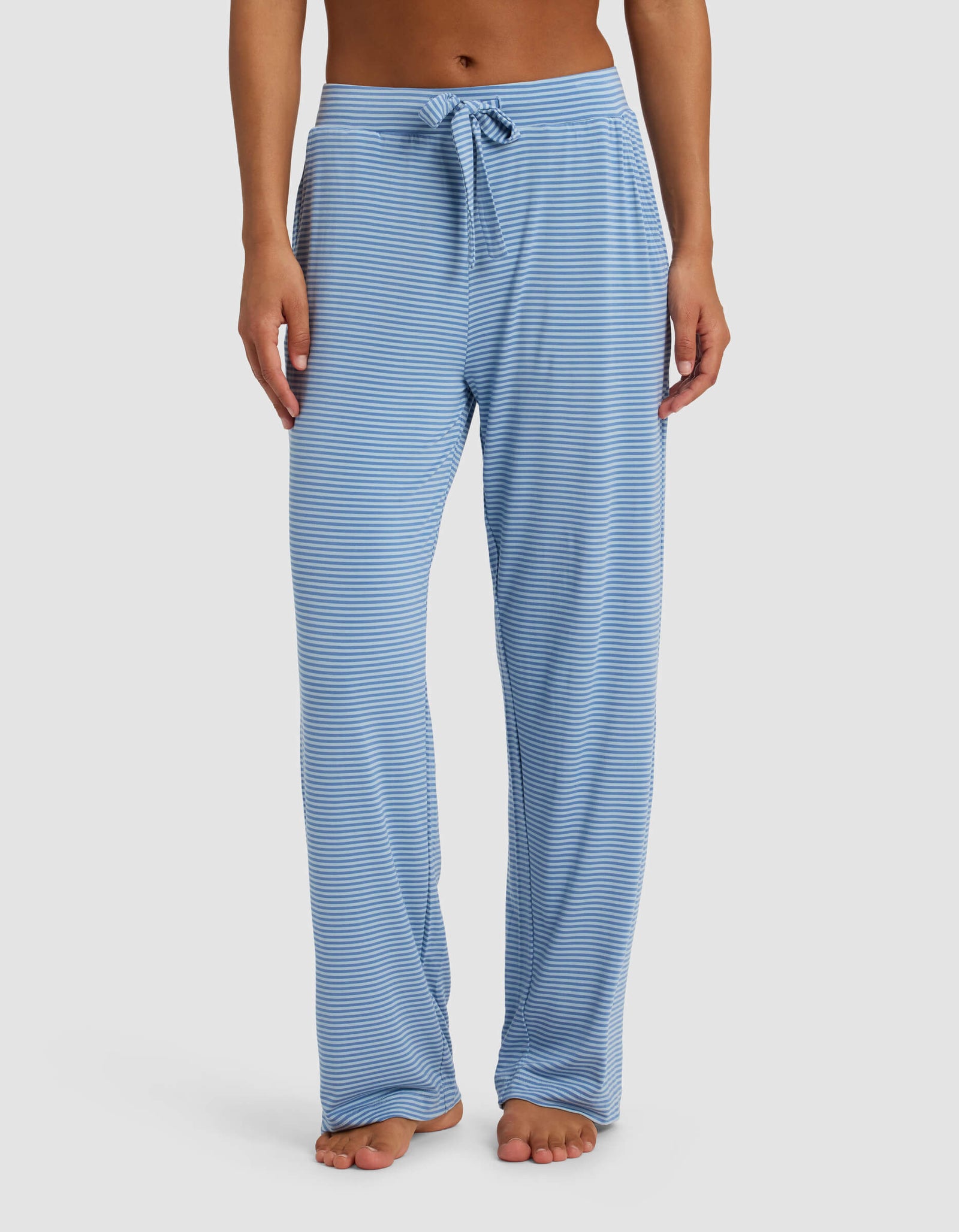 A person wears Cozy Earth's Women's Bamboo Stretch-Knit Pant—loose-fitting, light blue and white striped with a drawstring waist—standing barefoot against a plain white background.