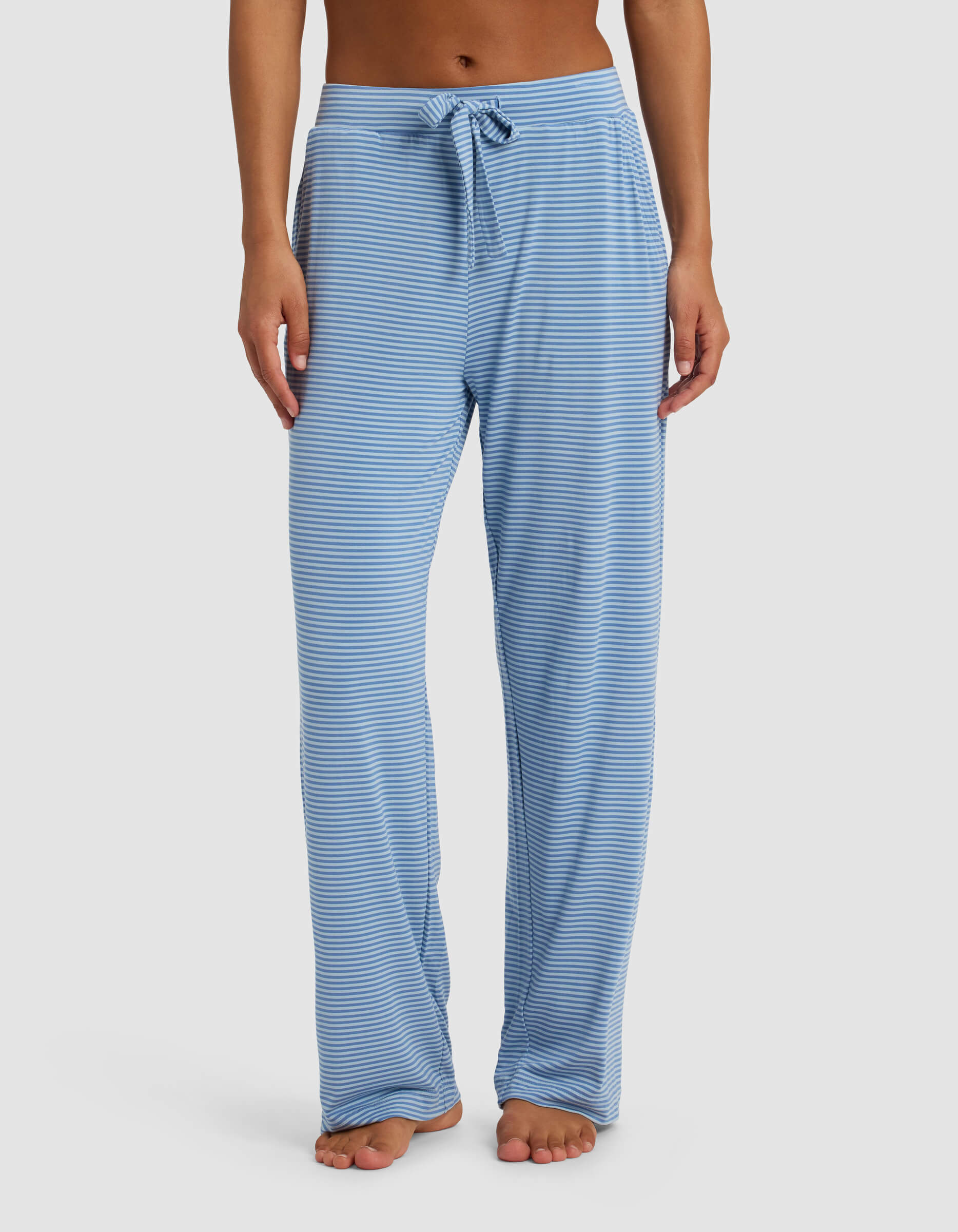 A person wears Cozy Earth's Women's Bamboo Stretch-Knit Pant—loose-fitting, light blue and white striped with a drawstring waist—standing barefoot against a plain white background.|Color:Sailor Mini Stripe