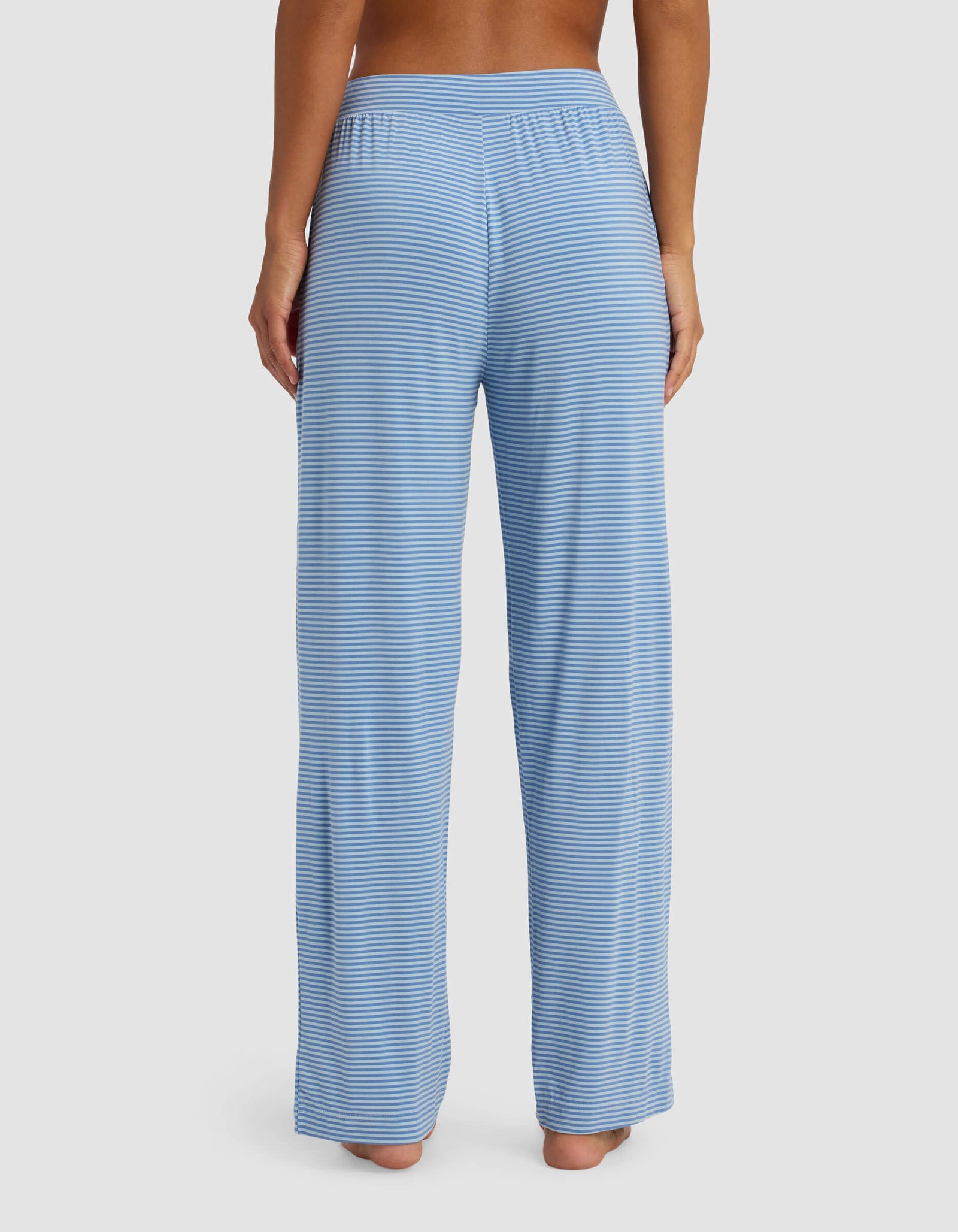A woman is seen from the back, barefoot, wearing Cozy Earth's Women's Bamboo Stretch-Knit Pant in light blue and white stripes, standing against a plain white background.
