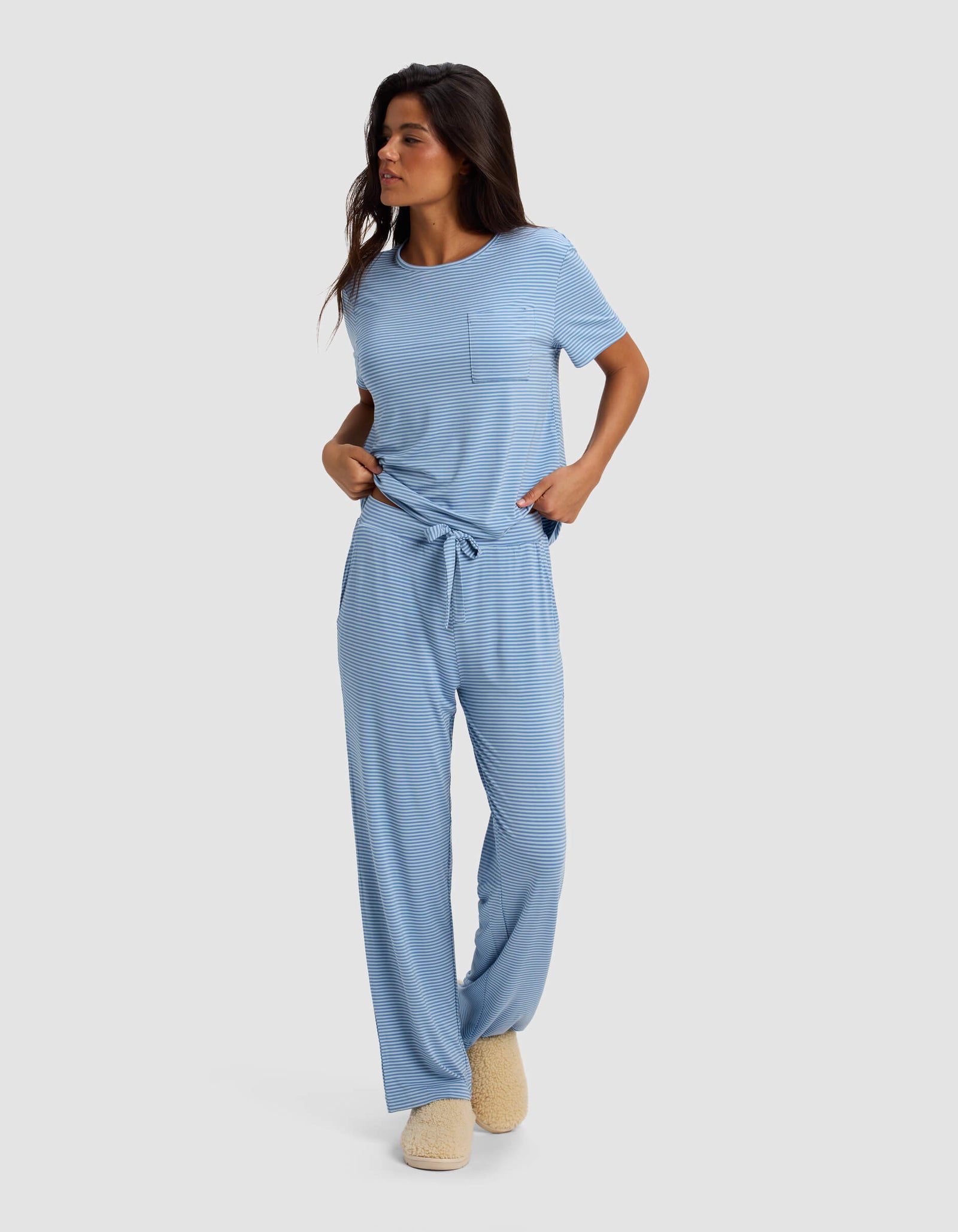 A woman stands against a light gray background wearing Cozy Earth Women's Bamboo Stretch-Knit Pant pajamas in light blue stripes with short sleeves, a front pocket, drawstring waist, and fuzzy beige slippers.
