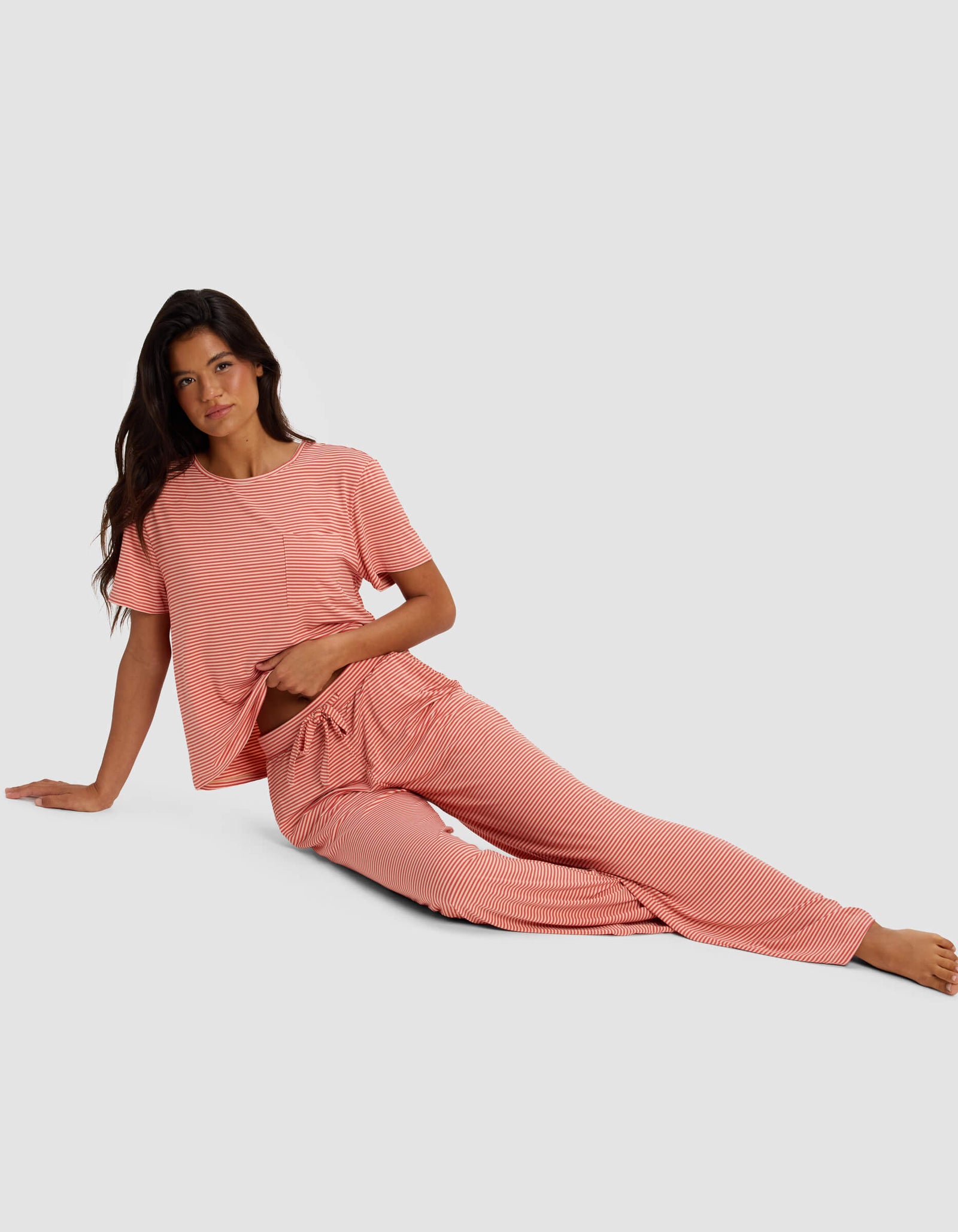 A woman with long dark hair models Cozy Earth Women’s Bamboo Stretch-Knit Pants in pink and white stripes, sitting on the floor with one arm propped behind her and legs extended against a plain light gray background.