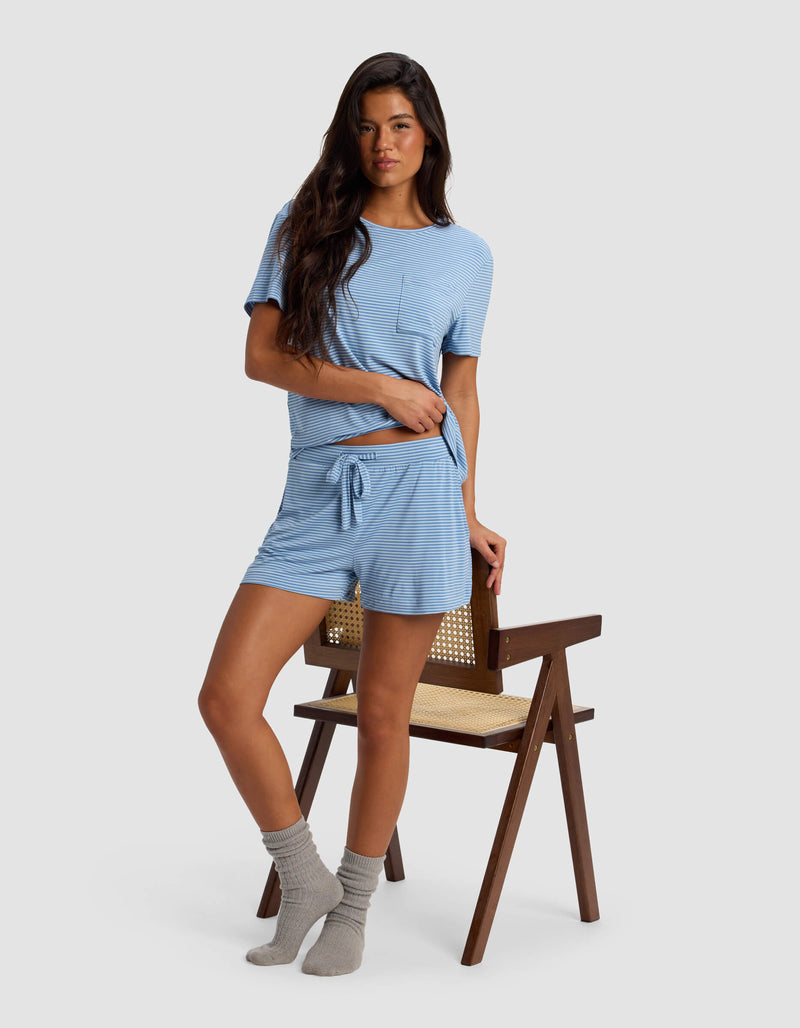 A woman with long dark hair and a relaxed expression stands next to a wooden chair, wearing Cozy Earth's Women's Bamboo Stretch-Knit Shorts with a light blue striped t-shirt and gray socks, set against a plain light gray background.