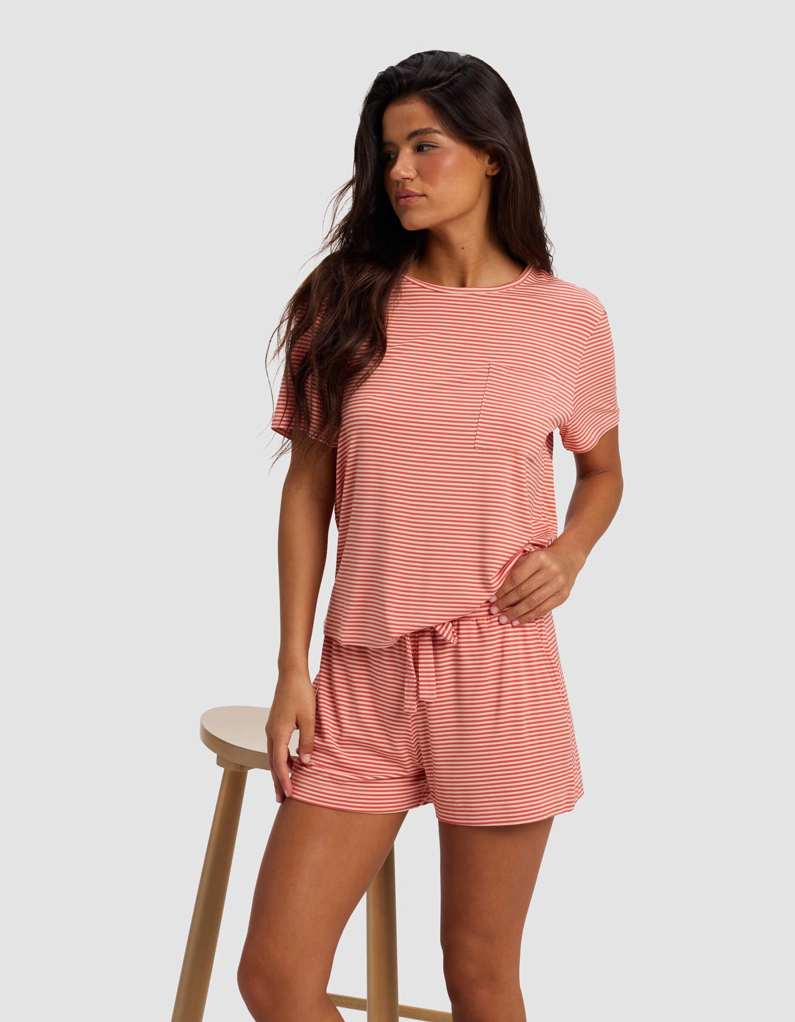 A woman stands beside a wooden stool, wearing Cozy Earth’s Women's Bamboo Stretch-Knit Shorts as part of a pink and white striped short-sleeve pajama set, looking off to the side against a plain light gray background.