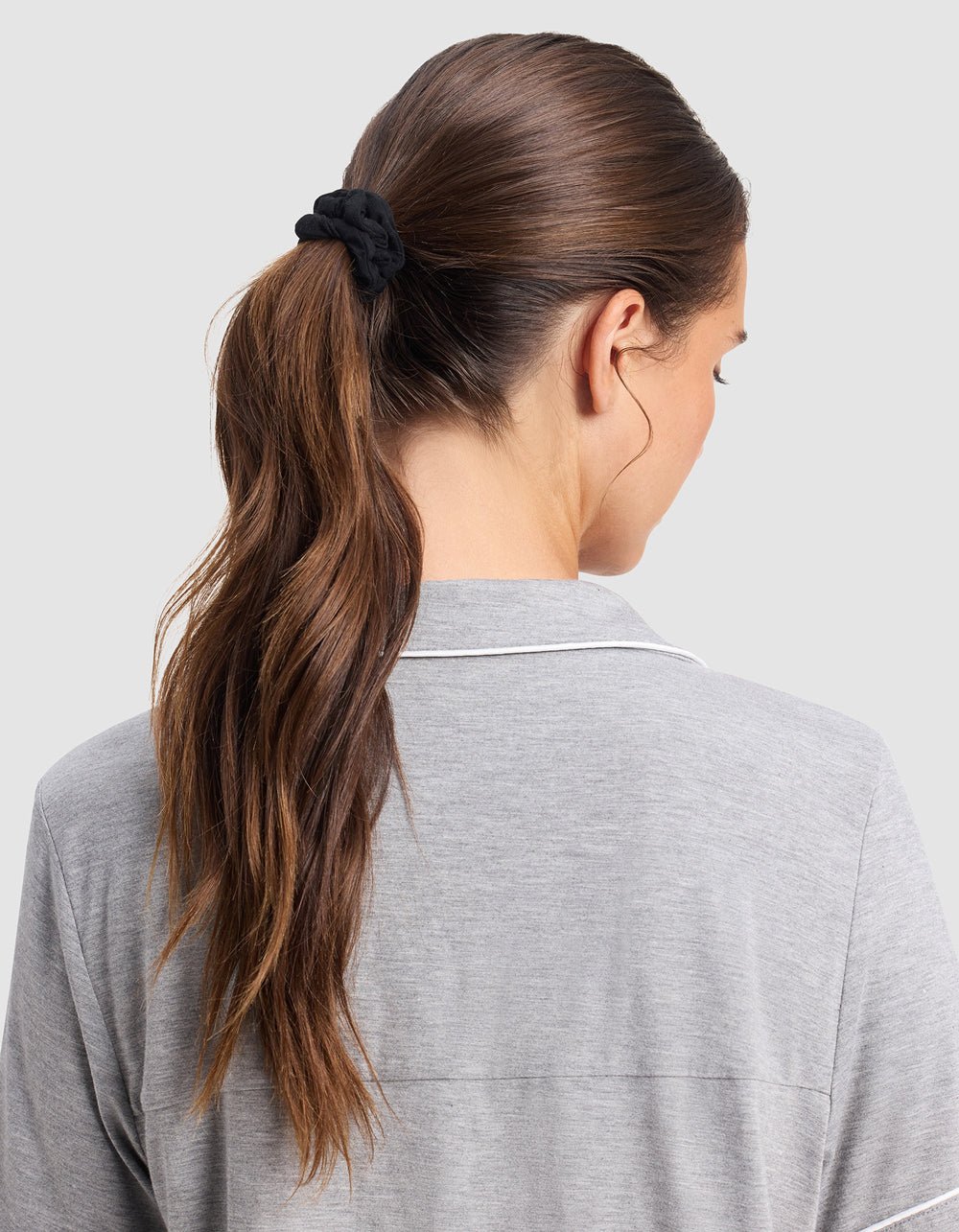 A woman with long brown hair tied in a ponytail with a black Cozy Earth Bamboo Stretch Knit Skinny Scrunchie, wearing a light gray collared shirt, is seen from the back against a plain light background.