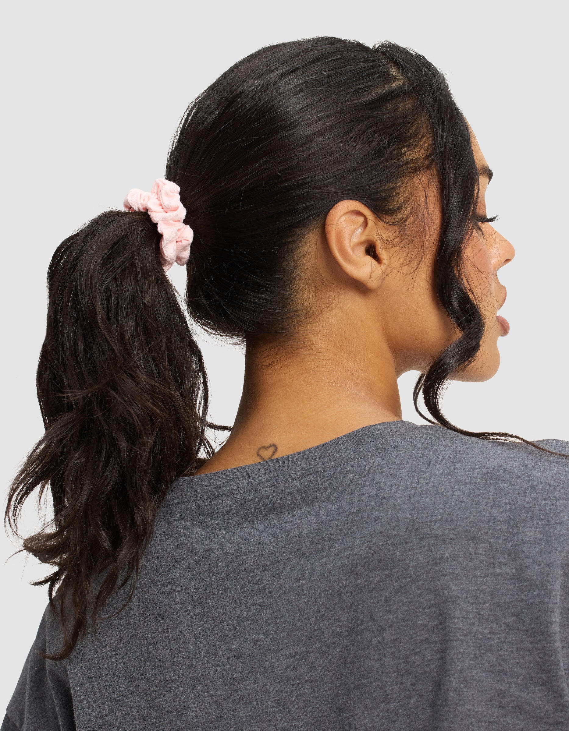 A woman with dark hair in a ponytail, secured by a Cozy Earth Bamboo Stretch Knit Skinny Scrunchie in light pink, wears a gray shirt and faces away from the camera, revealing a small heart tattoo at her nape. 