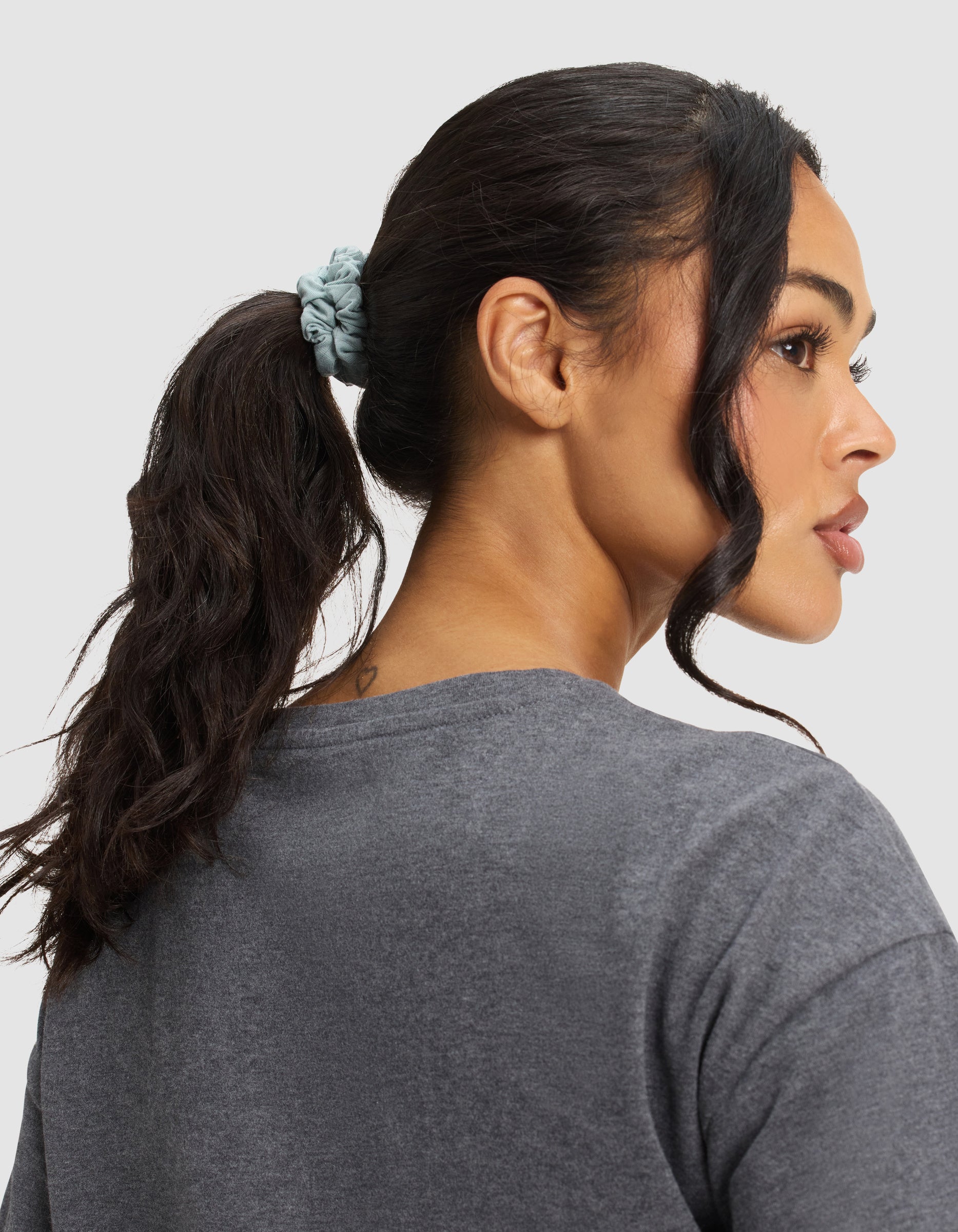 Woman with long dark hair in a ponytail held by a light blue Cozy Earth Bamboo Stretch Knit Skinny Scrunchie, wearing a grey t-shirt and showing a small heart tattoo on the back of her neck, looks to the right. 
