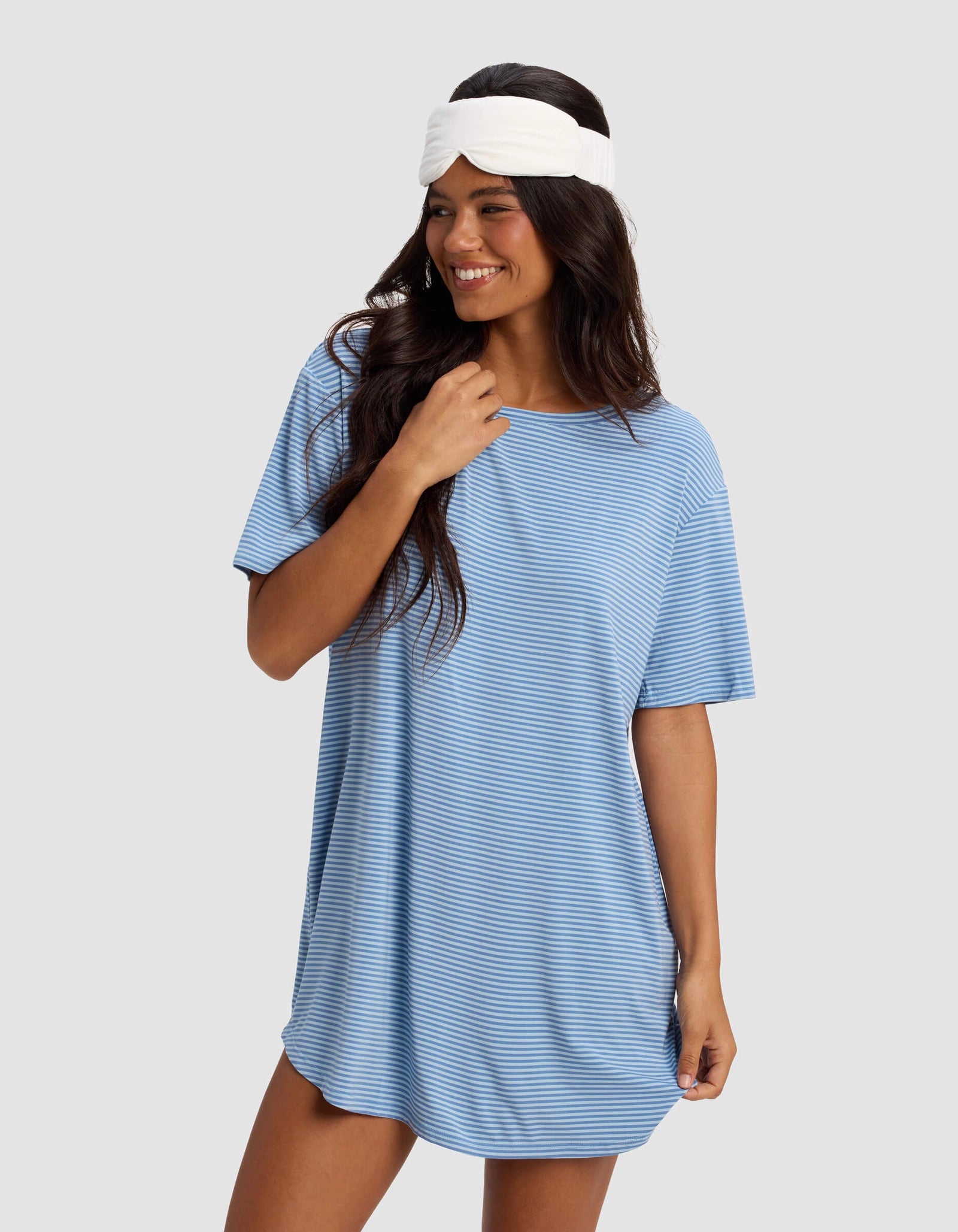 A woman in a Cozy Earth Women's Bamboo Stretch Knit Sleep Dress, featuring loose short sleeves and blue and white stripes, smiles while looking to the side against a plain light gray background.