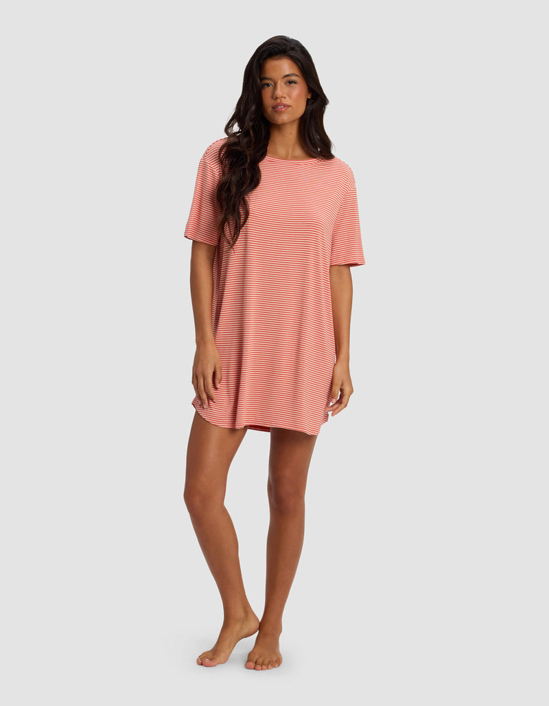 A woman stands barefoot in a Cozy Earth Women's Bamboo Stretch Knit Sleep Dress, featuring loose-fitting short sleeves and pink and white stripes, set against a plain light gray background.