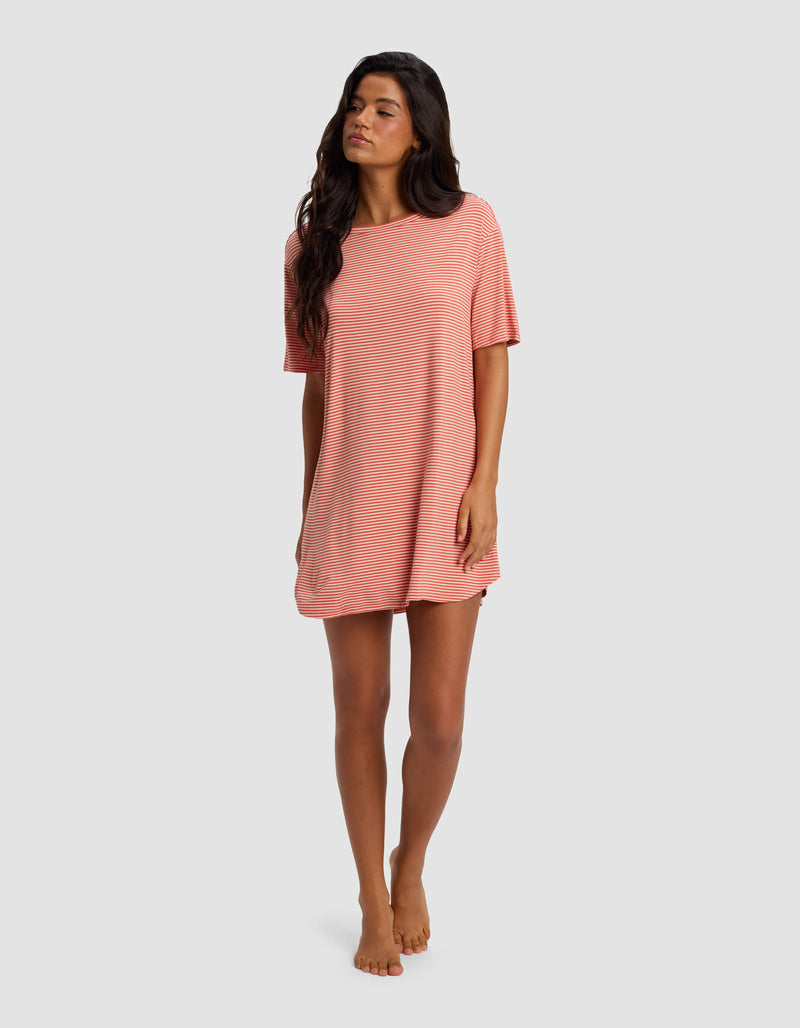 A woman with long dark hair stands barefoot against a light gray background, wearing the Cozy Earth Women's Bamboo Stretch Knit Sleep Dress—a loose-fitting, short-sleeved style in light pink and white stripes.