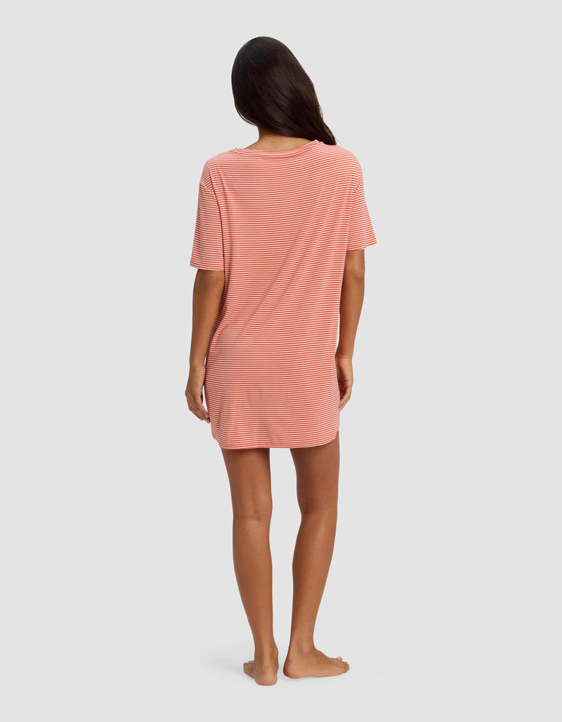 A woman stands barefoot facing away, wearing the Cozy Earth Women's Bamboo Stretch Knit Sleep Dress in pink and white stripes. The loose, short-sleeved dress falls above her knees. Her long dark hair is down against a light gray background.