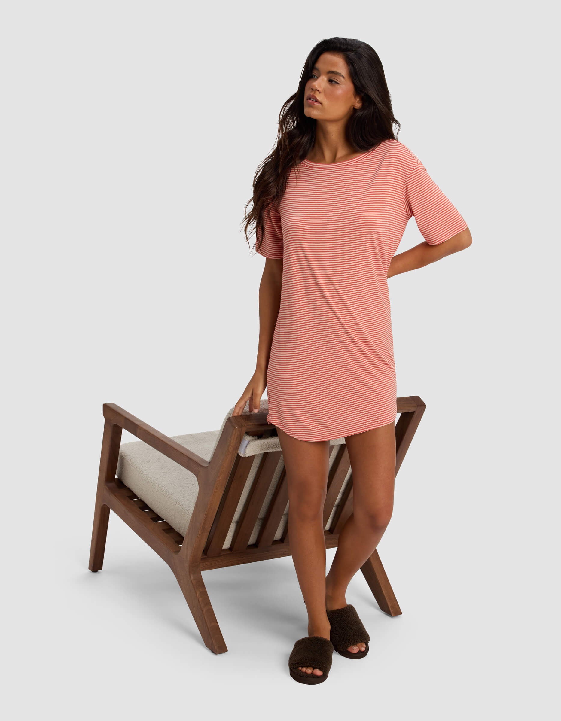 A woman in the Cozy Earth Women's Bamboo Stretch Knit Sleep Dress stands beside a wooden chair with a beige cushion, wearing dark slippers and looking to the side against a plain background.