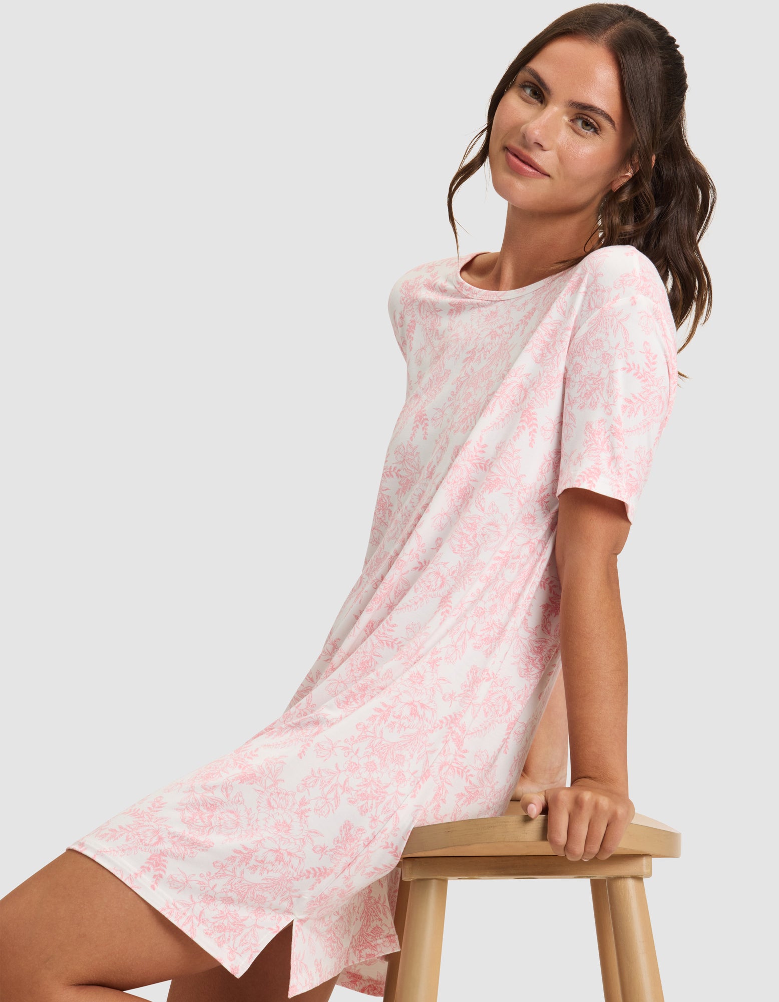 A woman with long brown hair sits sideways on a wooden stool, gently smiling at the camera while wearing Cozy Earth's Women's Bamboo Stretch-Knit Sleep Dress. 