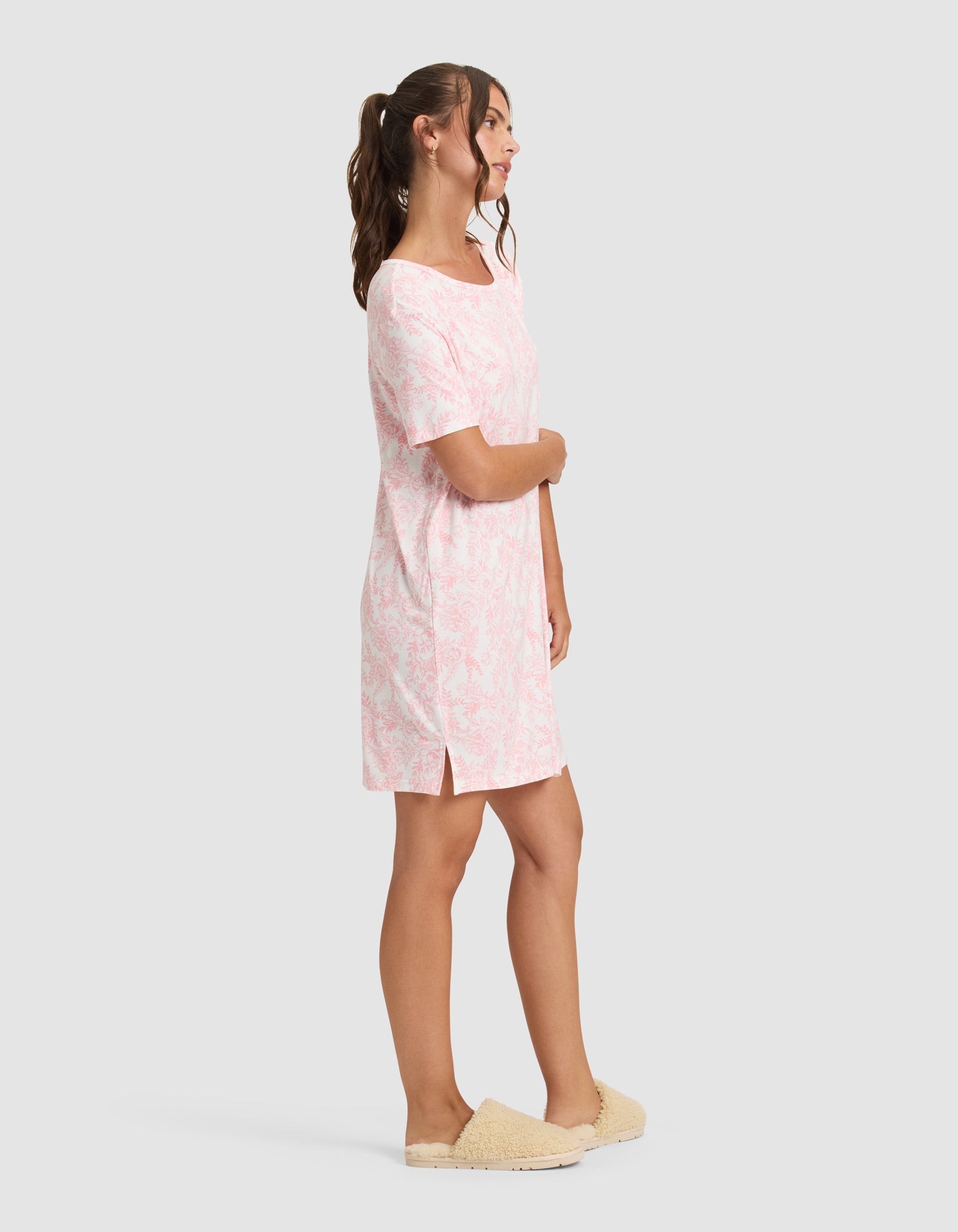 A woman stands in profile wearing the Cozy Earth Women's Bamboo Stretch-Knit Sleep Dress, featuring light pink and white patterns with side slits, paired with beige fuzzy slippers. Her hair is in a ponytail against a plain light gray background. 