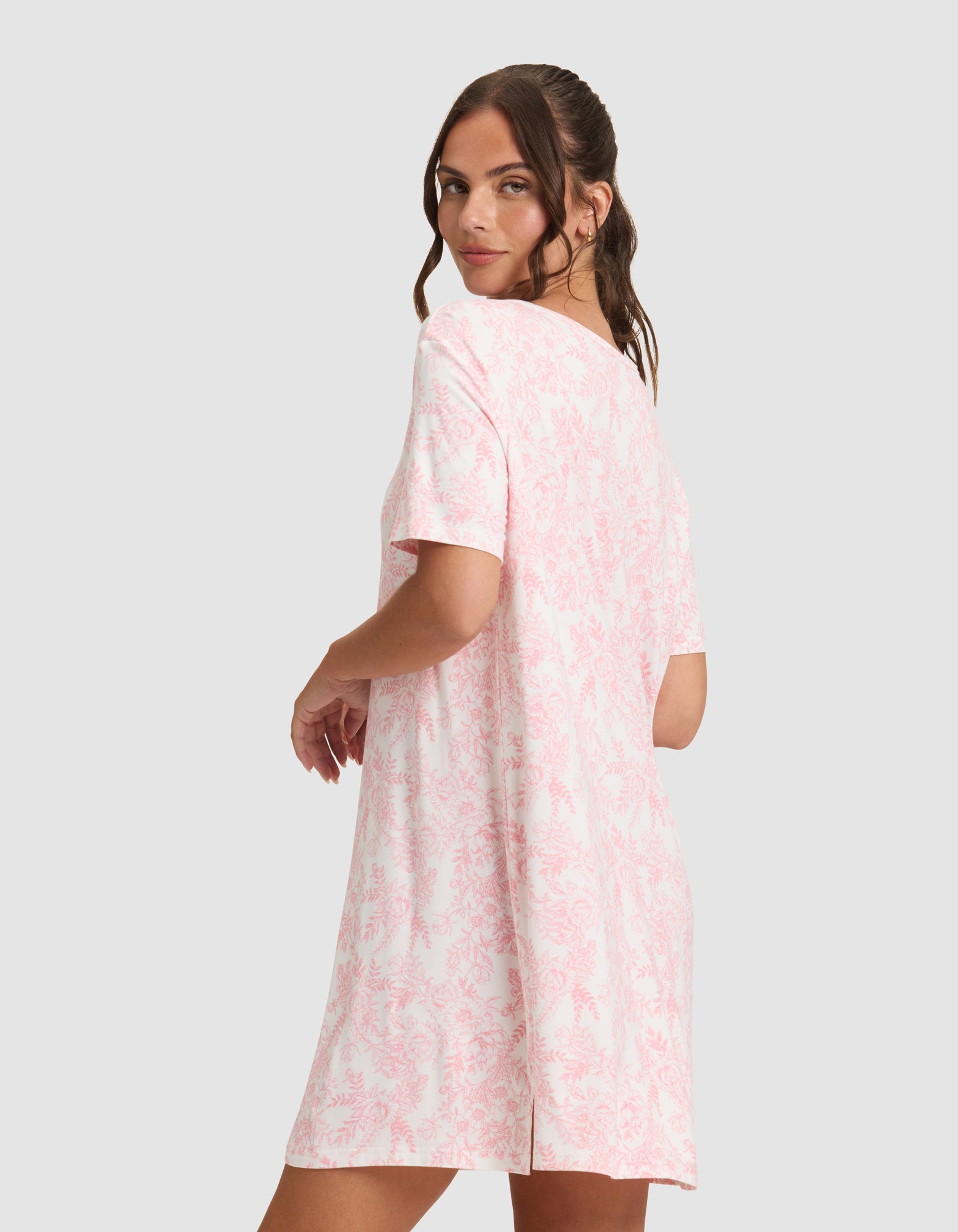 A woman with long brown hair models the Cozy Earth Women's Bamboo Stretch-Knit Sleep Dress—a loose, short-sleeved, knee-length pink and white patterned dress—standing sideways against a plain light background. 