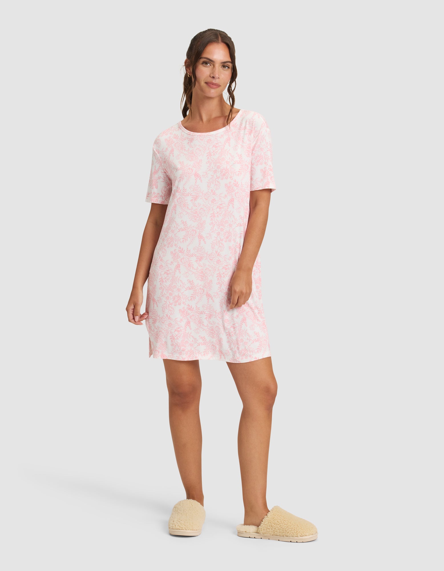 A woman with long, wavy hair and a relaxed expression wears the Cozy Earth Women's Bamboo Stretch-Knit Sleep Dress against a plain background, paired with tan fluffy slippers. 