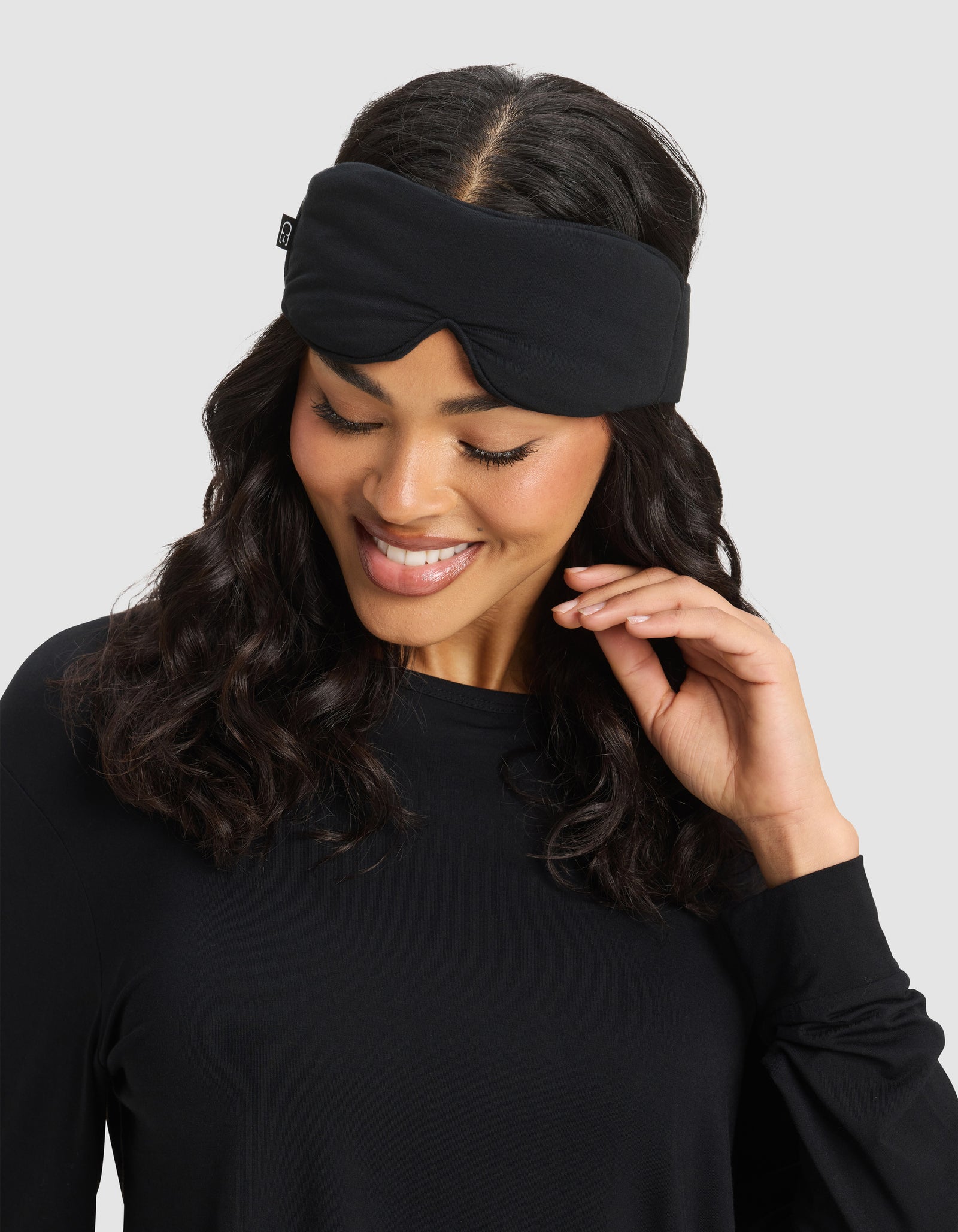 A woman with wavy dark hair smiles while wearing the Cozy Earth Bamboo Stretch-Knit Sleep Mask pushed up on her forehead and a black long-sleeve top, touching her hair against a plain light gray background.