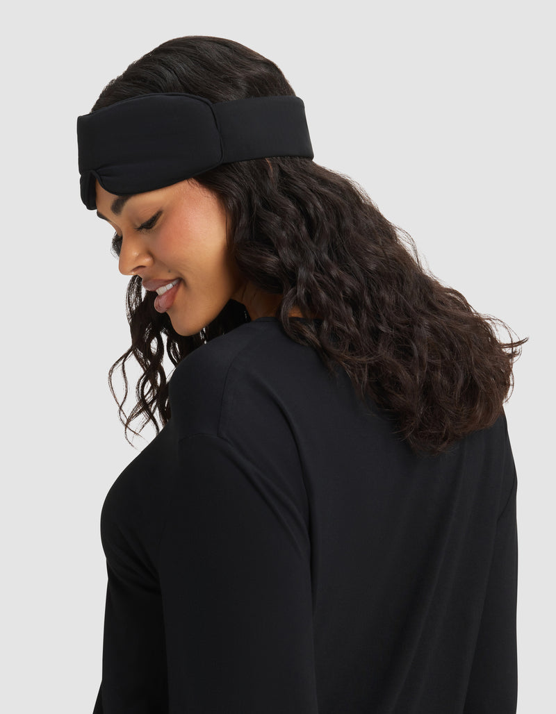 A woman with wavy dark hair smiles as she looks down, wearing the Cozy Earth Bamboo Stretch-Knit Sleep Mask against a light gray background.