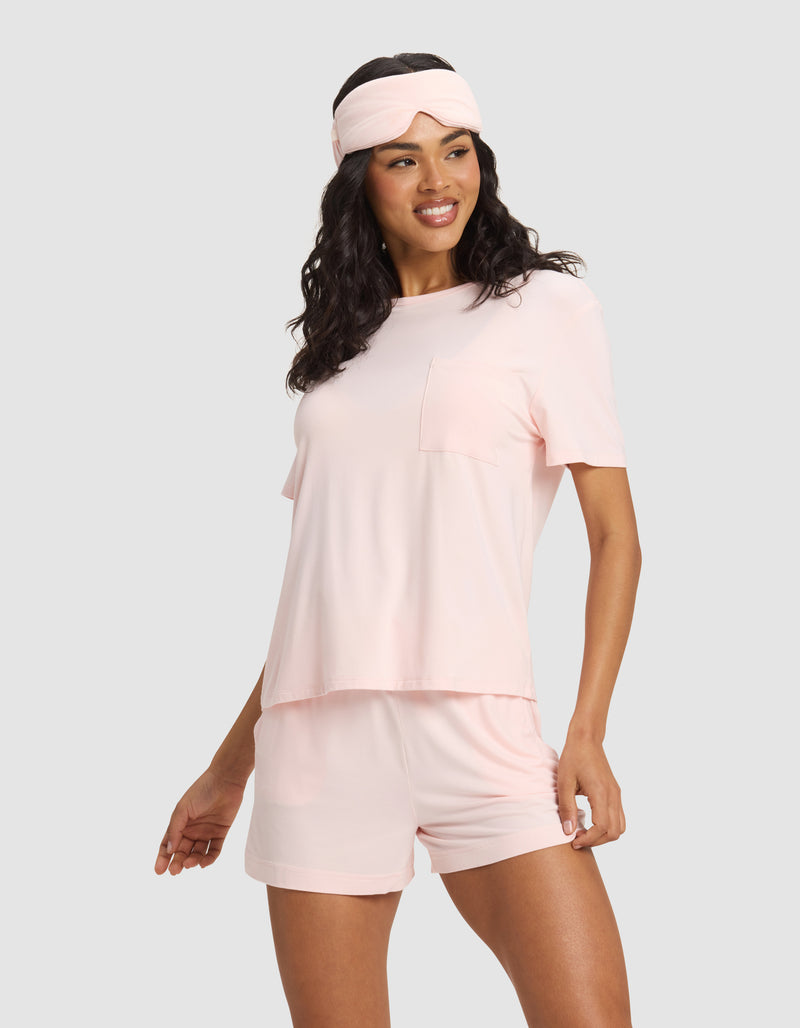 A woman stands smiling against a light gray background wearing a light pink pajama set with shorts and a Cozy Earth Bamboo Stretch-Knit Sleep Mask in matching pink on her head.