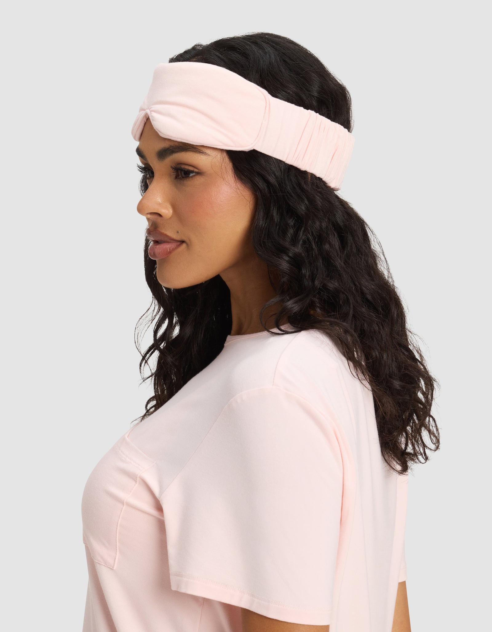 Woman with long, wavy dark hair wears a Cozy Earth Bamboo Stretch-Knit Sleep Mask, shown in profile against a plain light gray background. 