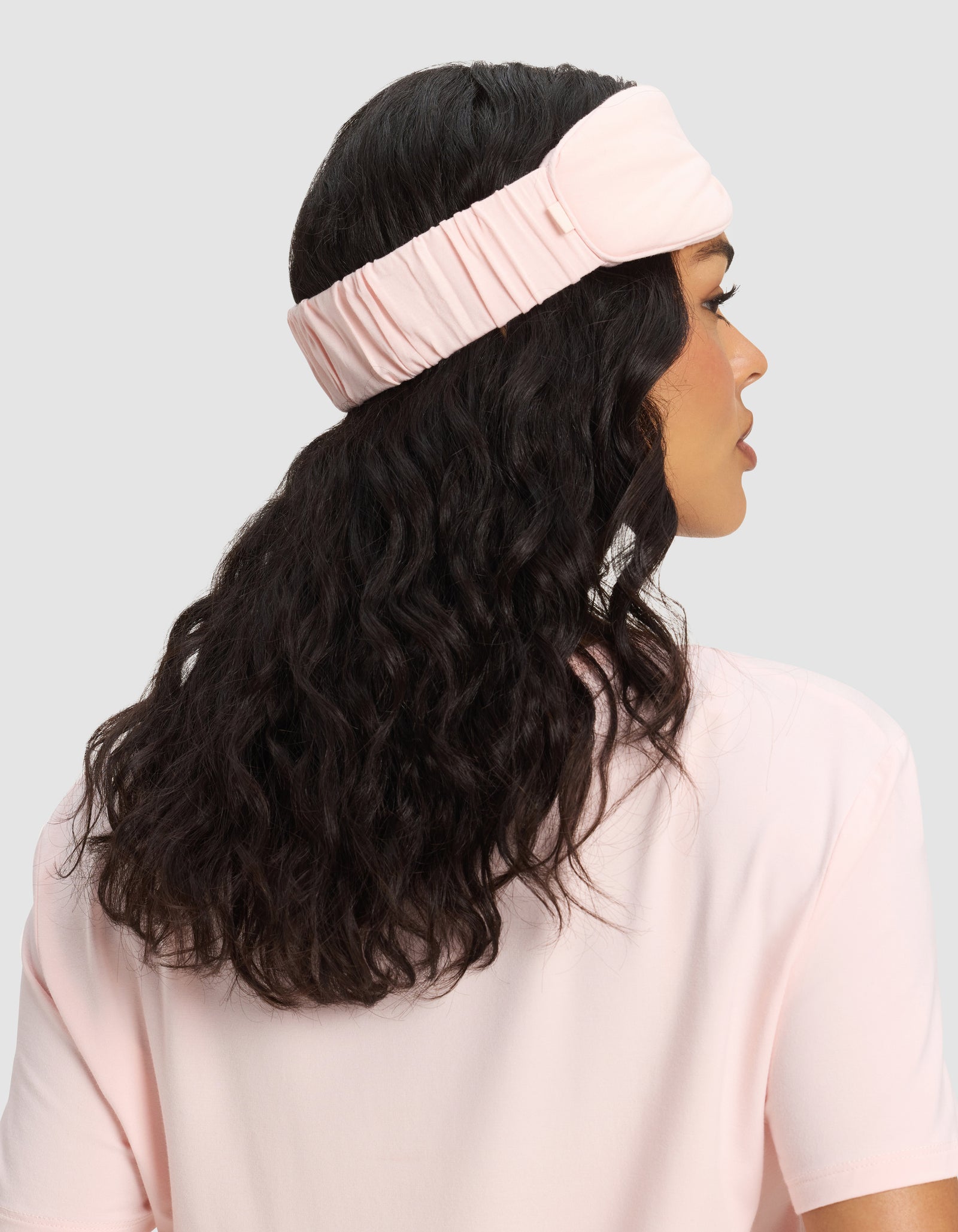A woman with wavy dark hair, wearing a Cozy Earth Bamboo Stretch-Knit Sleep Mask in light pink and a light pink top, is shown from the back against a plain gray background.