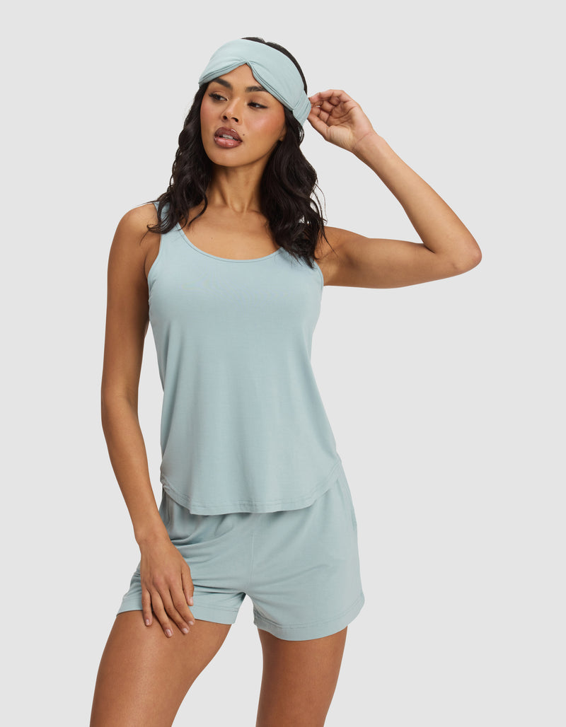 A woman poses against a light background, wearing a light blue sleeveless pajama set with matching shorts and the Cozy Earth Bamboo Stretch-Knit Sleep Mask, her hand gently touching the mask.