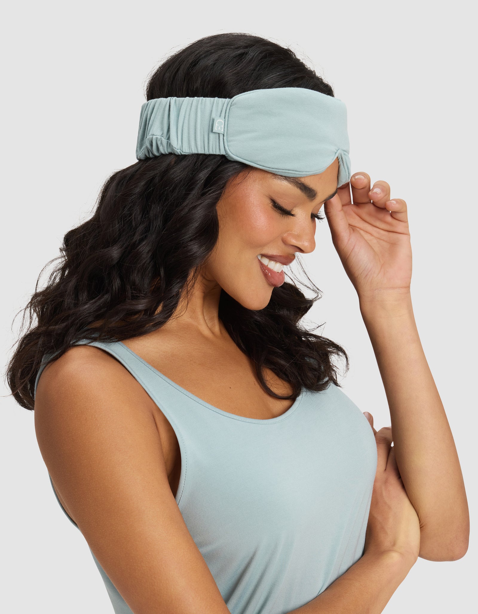 A woman with wavy dark hair smiles while wearing a Cozy Earth Bamboo Stretch-Knit Sleep Mask in light blue and a matching tank top, gently touching the mask with one hand. The background is plain light gray. 