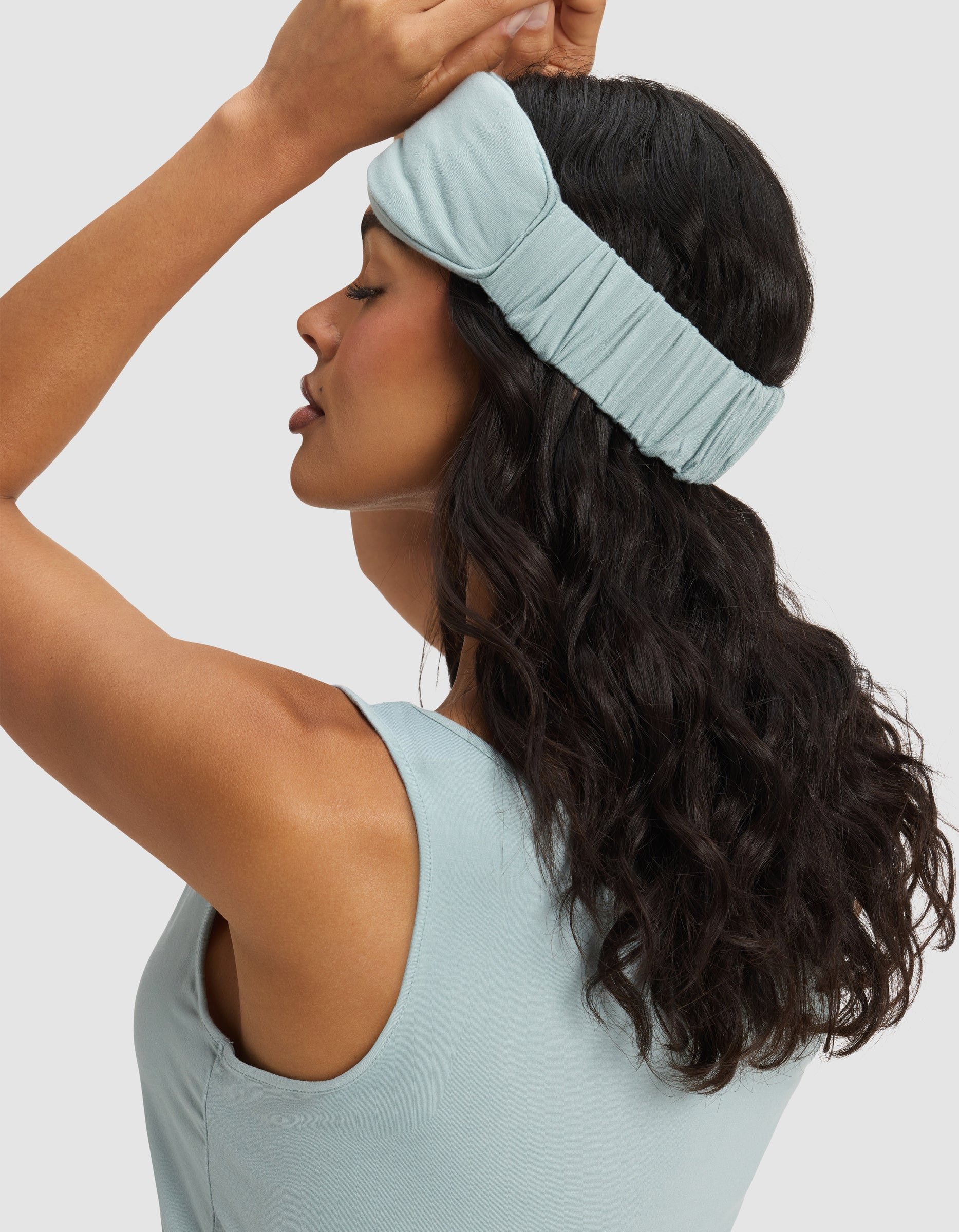 A woman with wavy dark hair wears a light blue tank and a Cozy Earth Bamboo Stretch-Knit Sleep Mask, pushed up on her forehead. She raises one hand to adjust the mask, her profile facing left against a light background.