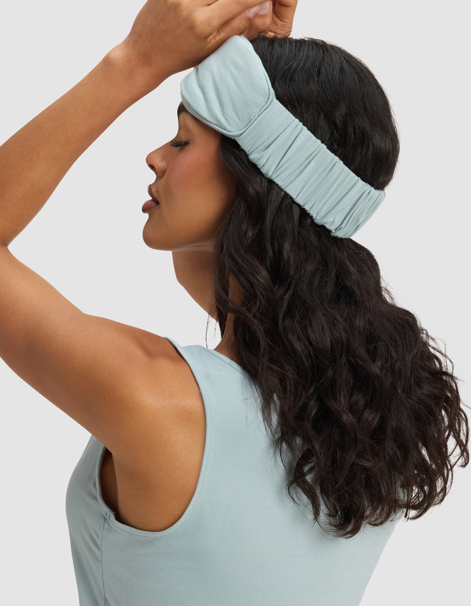 A woman with wavy dark hair wears a light blue tank and a Cozy Earth Bamboo Stretch-Knit Sleep Mask, pushed up on her forehead. She raises one hand to adjust the mask, her profile facing left against a light background.
