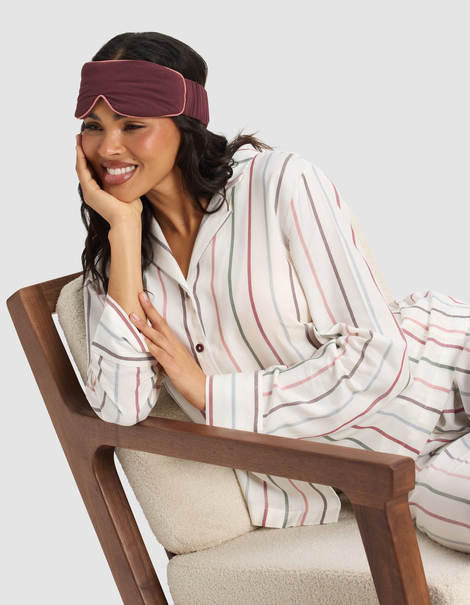 A woman smiles comfortably on a wooden armchair, wearing striped pajamas and a Cozy Earth Bamboo Stretch-Knit Sleep Mask with her head resting on one hand.